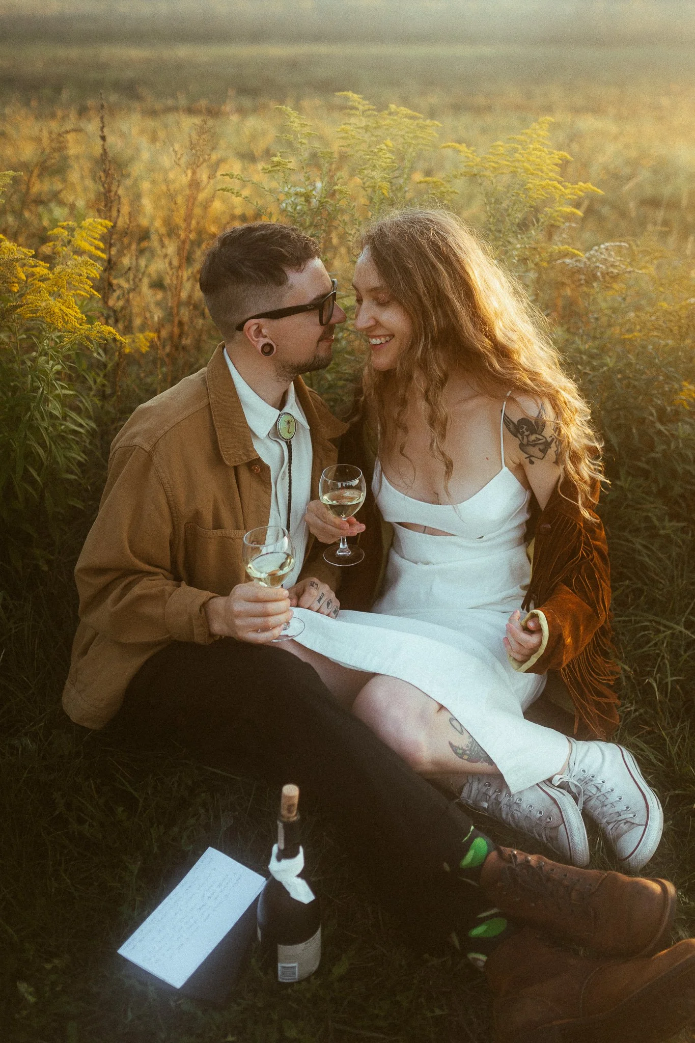 A happy couple sitting on the grass in a field during sunset, holding glasses of white wine, with a bottle and a note placed in front of them.