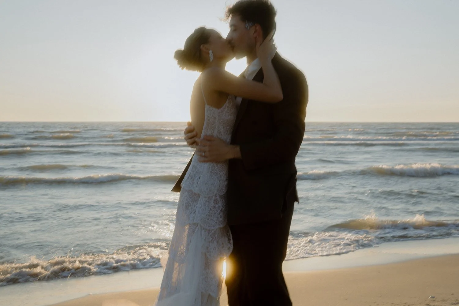 A couple in wedding attire kissing on a beach at sunset.