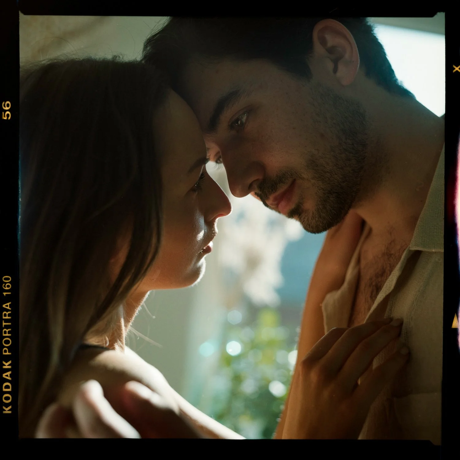 A close-up of a man and woman with their foreheads touching, looking into each other's eyes in an intimate moment indoors with sunlight streaming through a window.