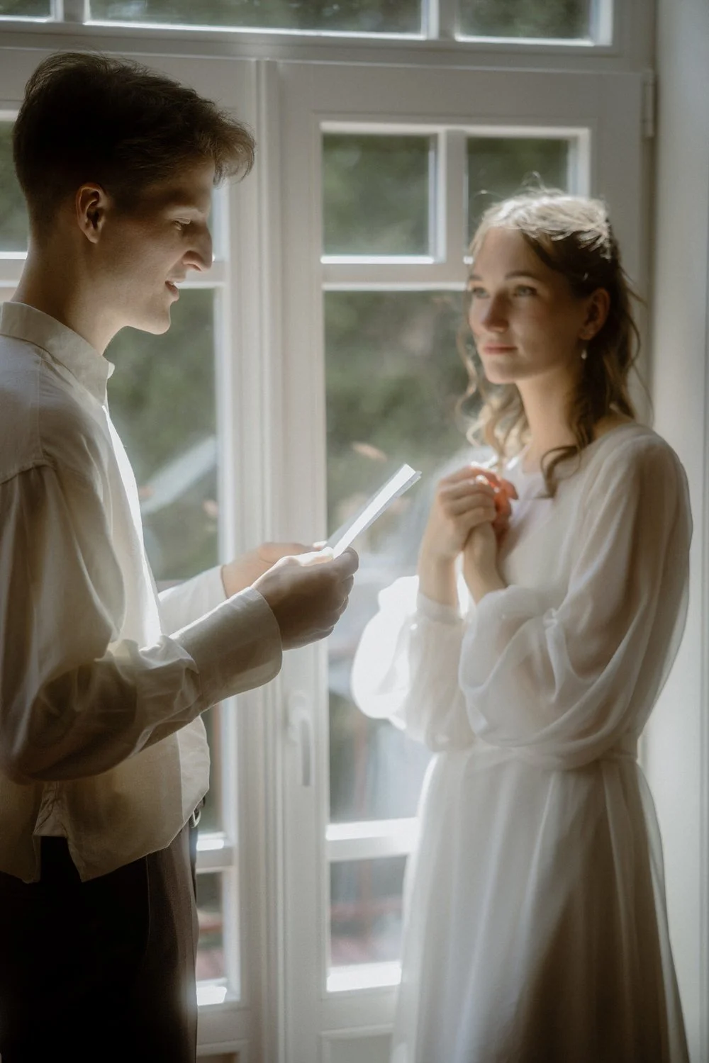 A young man reads a note or card to a young woman standing beside him by a window, with sunlight illuminating their faces. The woman looks at the man with a soft smile, hands clasped near her chest.