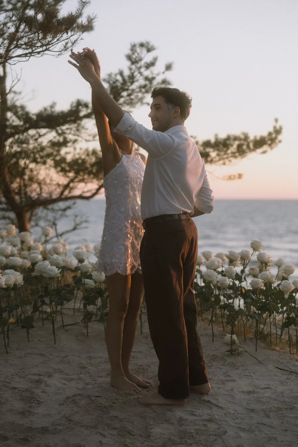 A couple dancing barefoot on the sand at sunset near the ocean, surrounded by white flowers and pine trees.