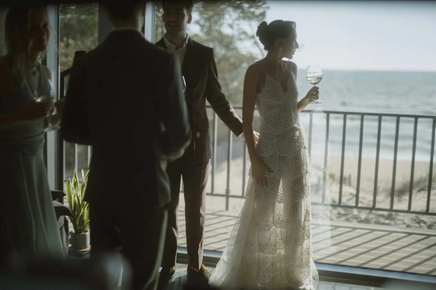 People at a wedding reception near a balcony overlooking the beach, with a woman in a lace dress holding a wine glass, and others engaged in conversation.