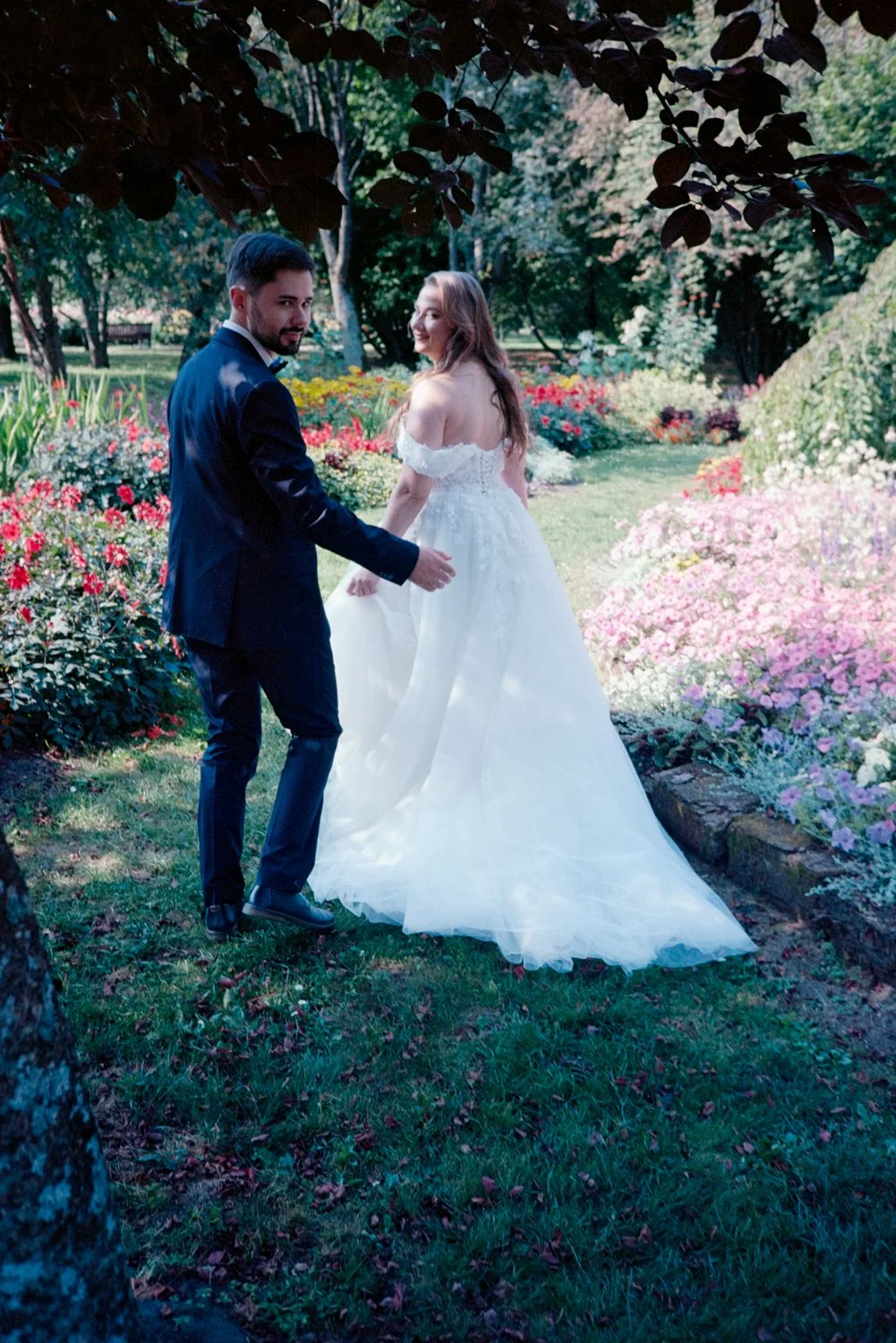 A bride and groom in wedding attire holding hands in a lush garden with colorful flowers and greenery, under a tree canopy.