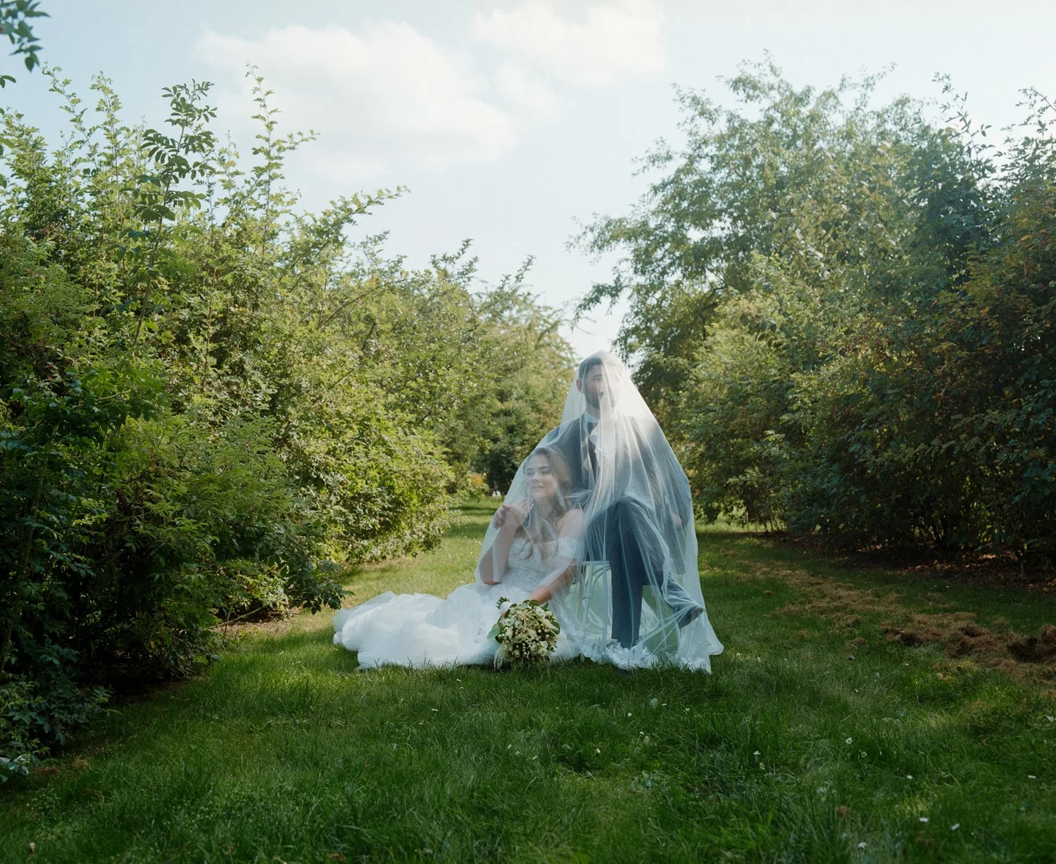 A bride and groom sit together outdoors on a grassy area, covered by a sheer veil, with greenery and trees in the background on a sunny day.