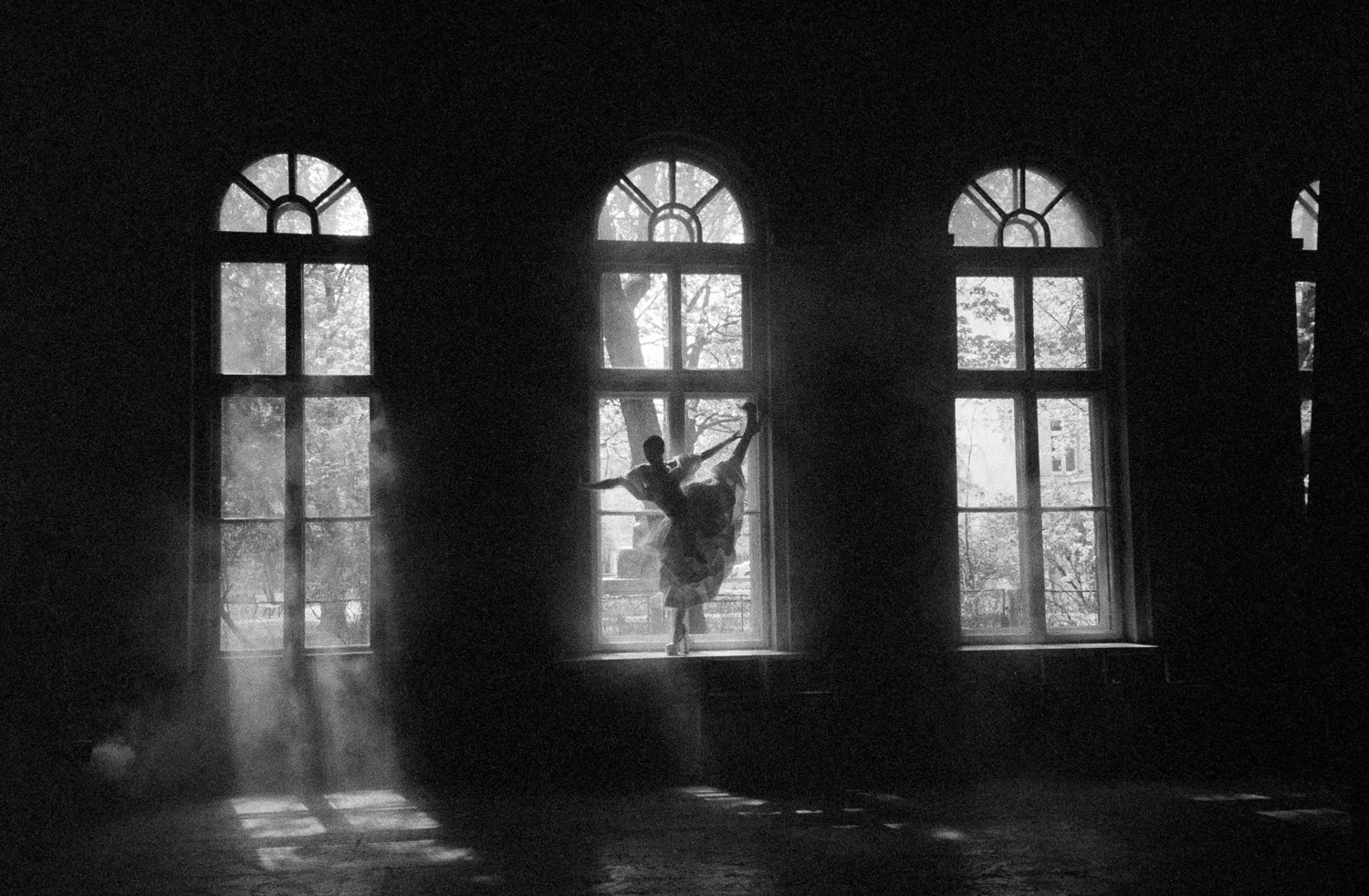 A ballet dancer practicing in front of large windows with sunlight streaming in, creating a silhouette effect.