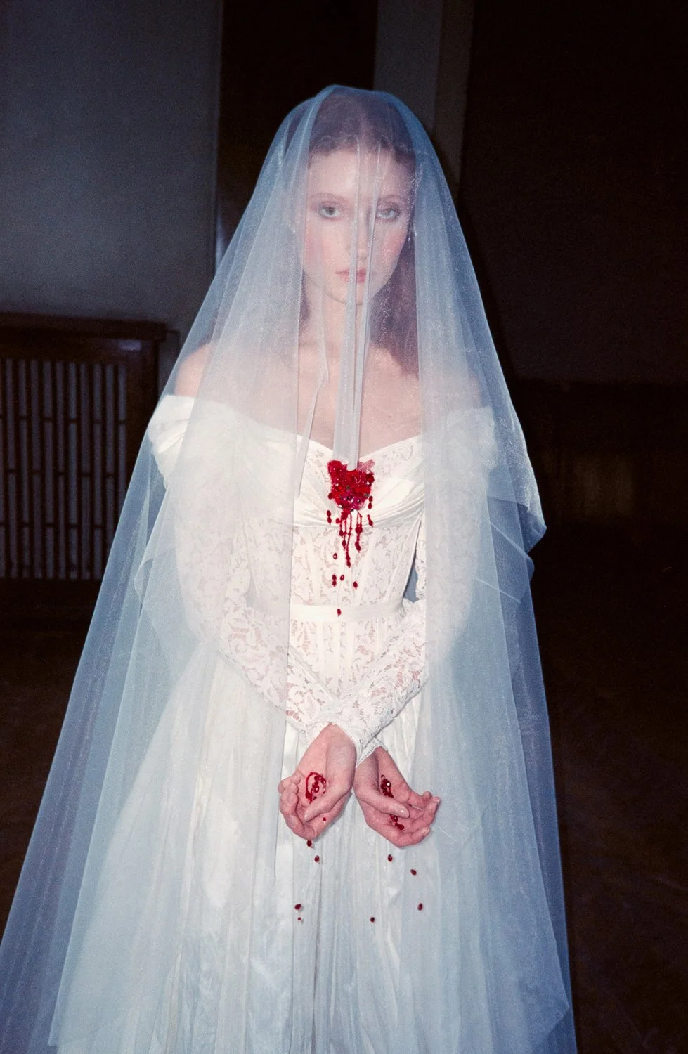 A woman dressed in a wedding gown with a veil, holding her hands with blood on her fingers, and a bloodstain on her chest, standing indoors.
