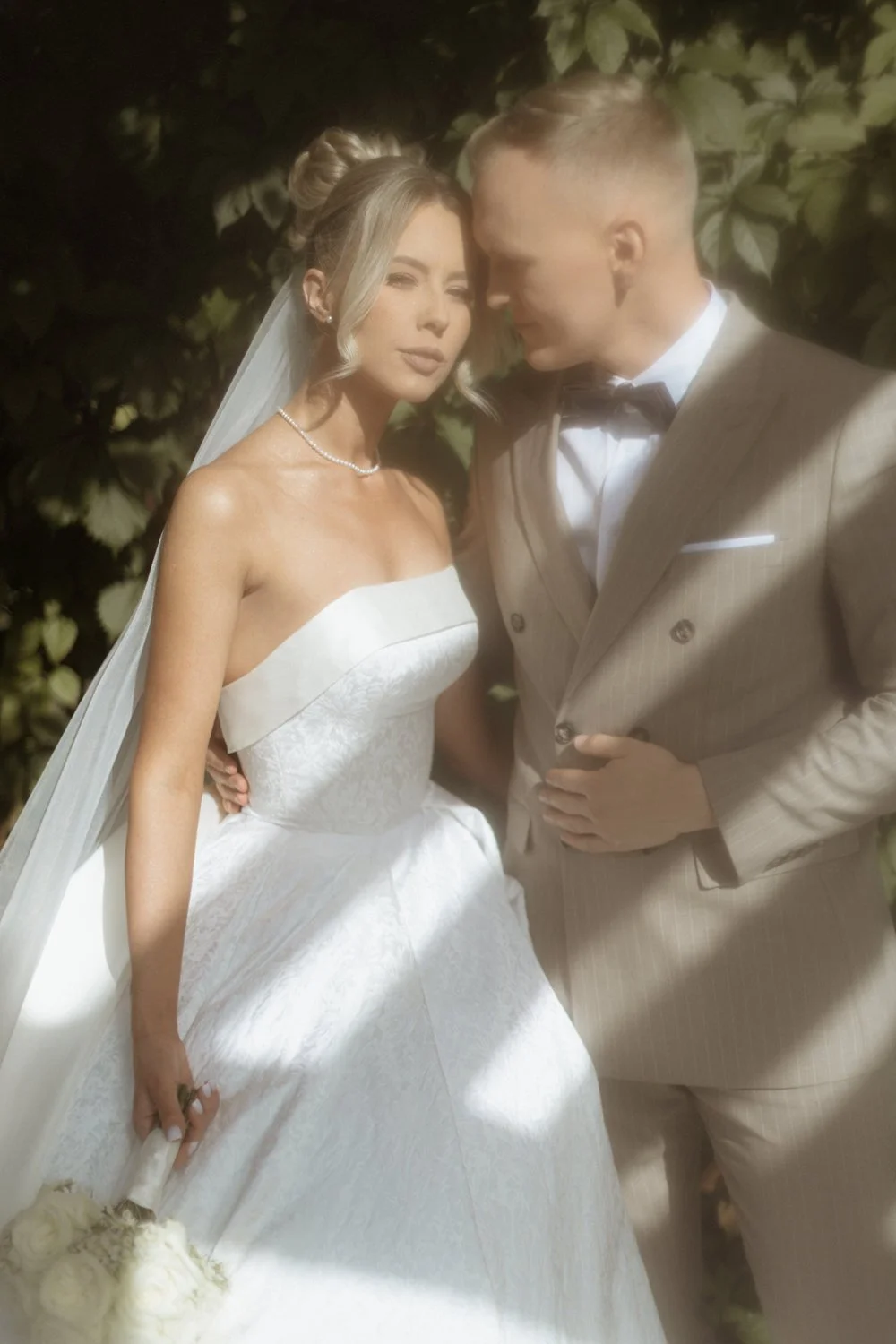 A bride and groom in wedding attire, standing close together outdoors with greenery in the background.