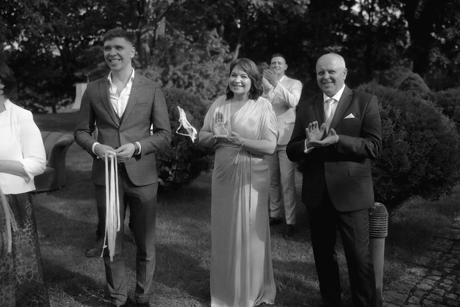 A black and white photo of people at a wedding or formal event outdoors, with three individuals in the foreground and others in the background, all smiling and making a gesture with their hands.