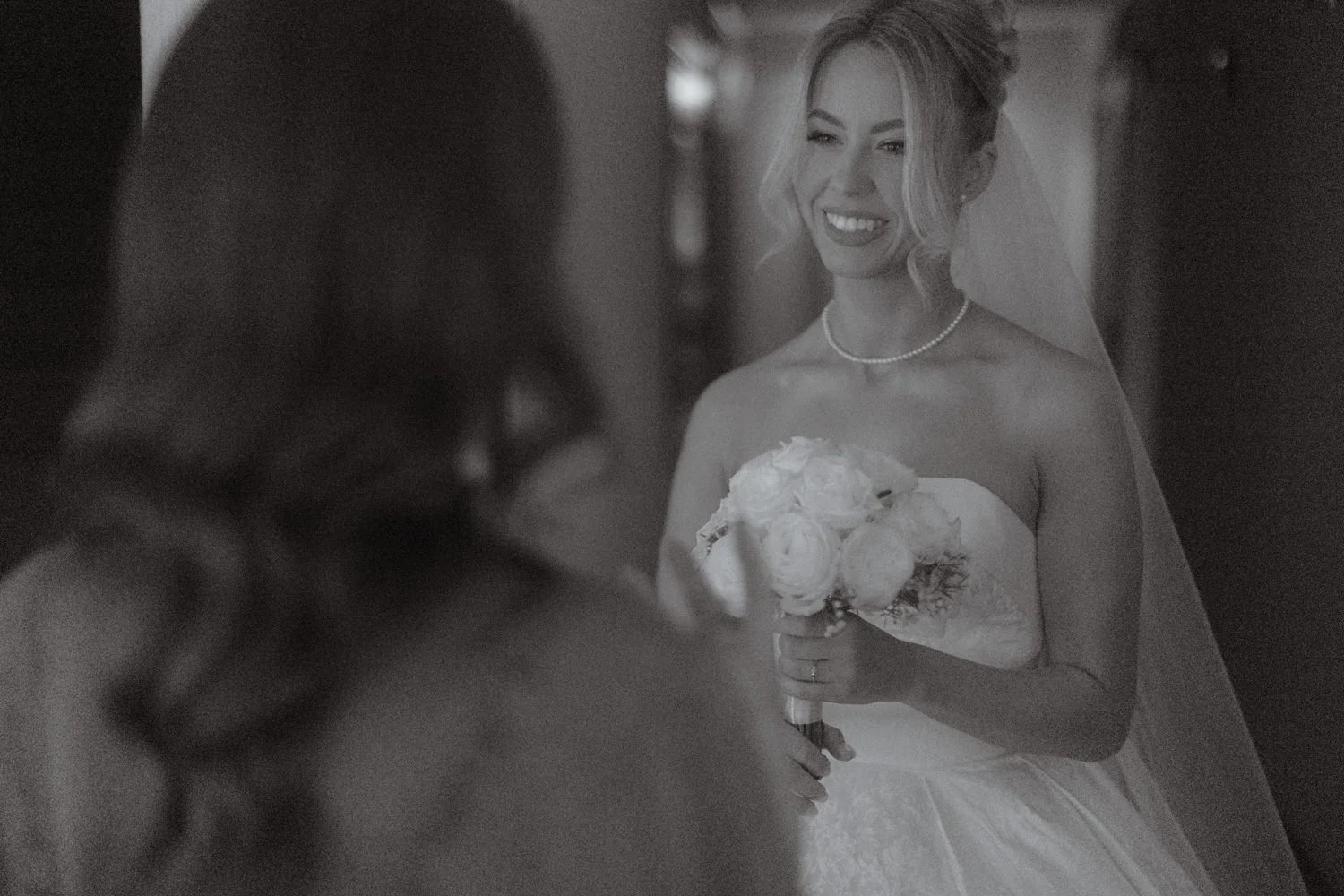 A bride in a wedding dress holding a bouquet of flowers and smiling at a bridesmaid.