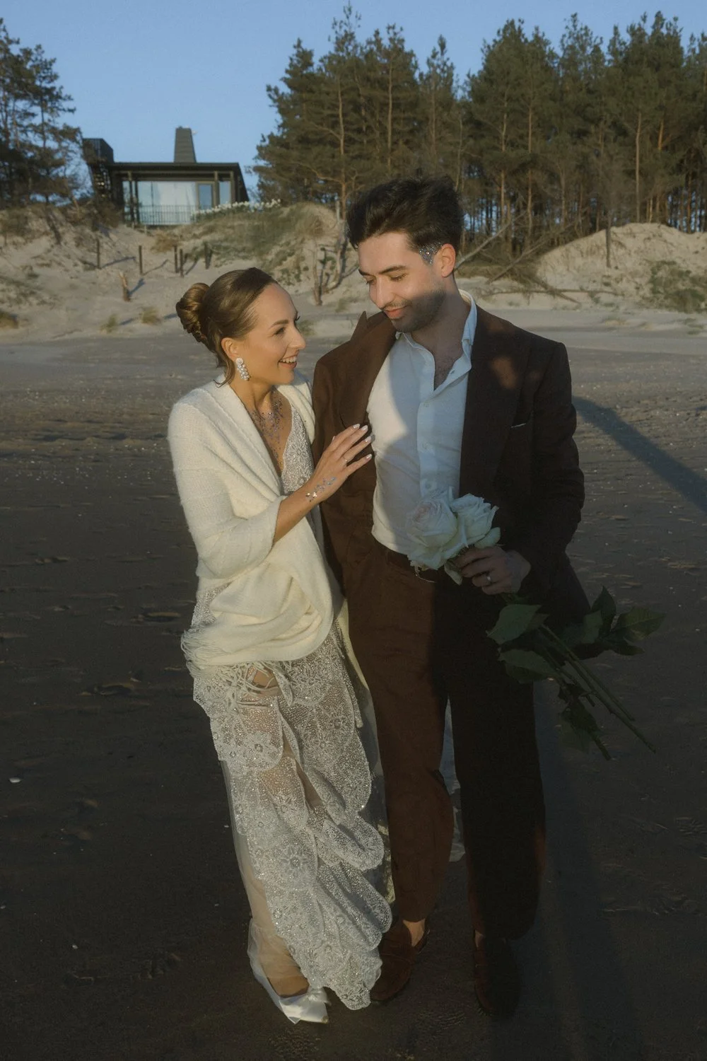 A couple dressed in wedding attire on the beach during sunset, holding a bouquet of white roses, with trees and a house in the background.