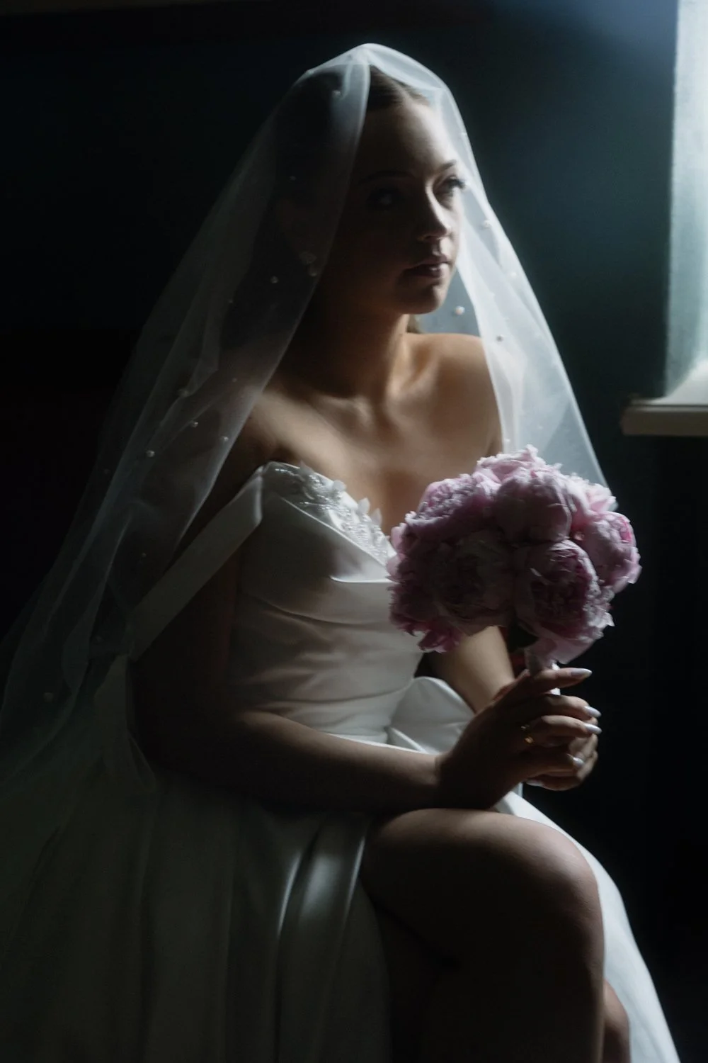 A bride wearing a white wedding dress and veil, holding a bouquet of pink flowers, sitting indoors near a window with light illuminating her face.