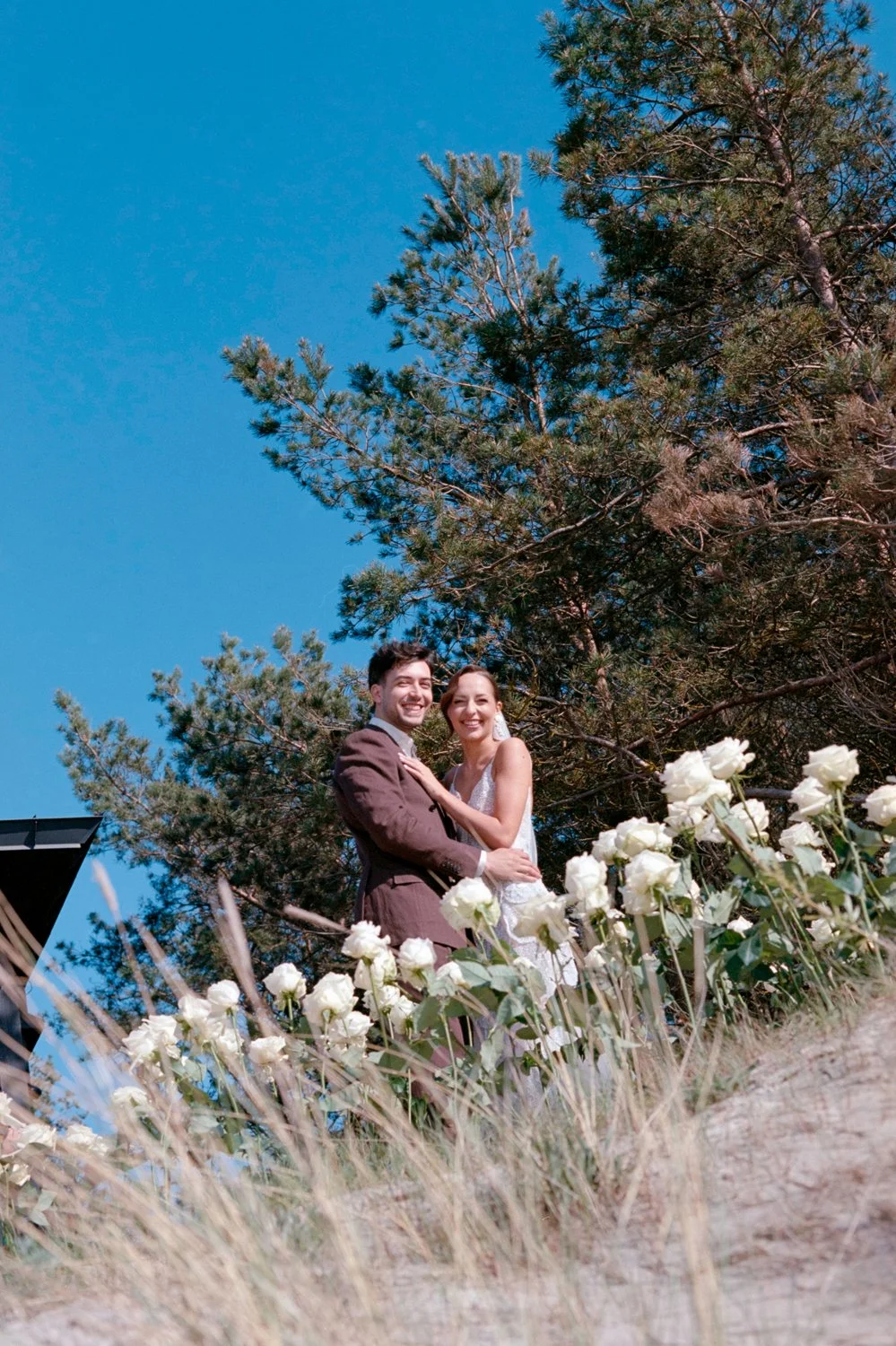 A happy couple dressed in wedding attire standing among white flowers, with large trees and a bright blue sky in the background.