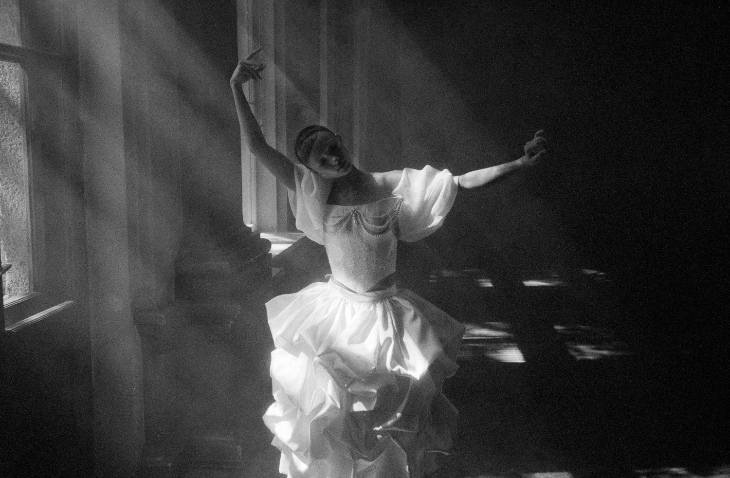 A woman dressed in a vintage, elegant gown dancing with arms outstretched in a dimly lit room with sunlight streaming through the windows.