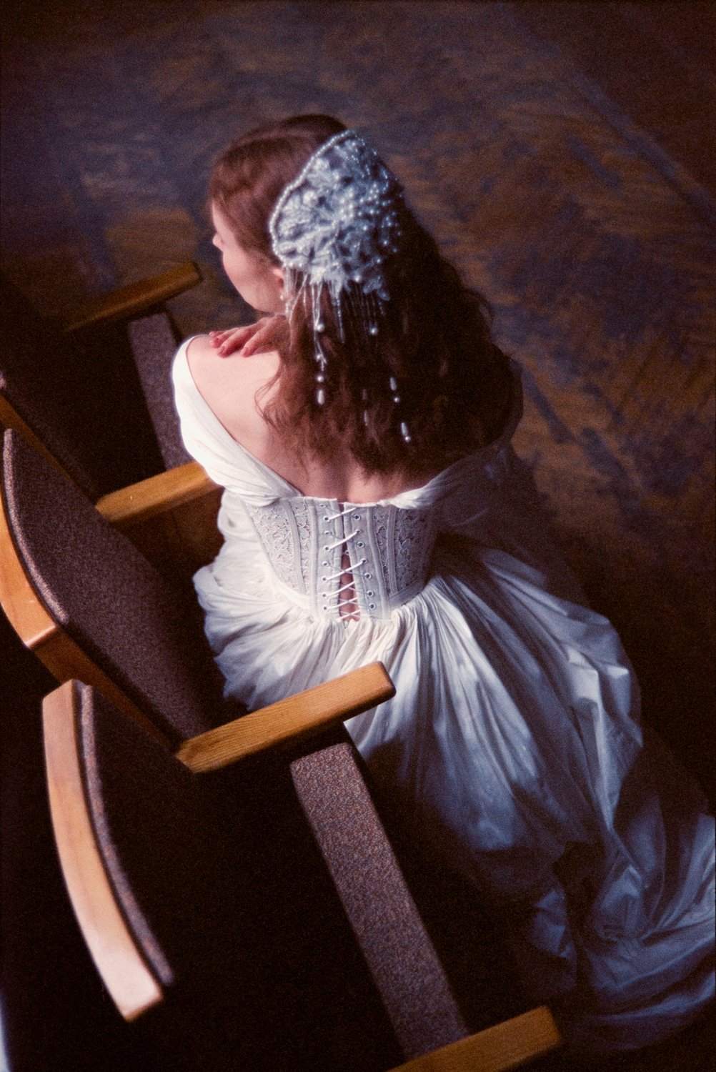 A woman dressed in a vintage-style gown and lace-up corset, sitting on a wooden pew with her back facing the camera. She has dark curly hair adorned with a decorative headpiece with beads and pearls.