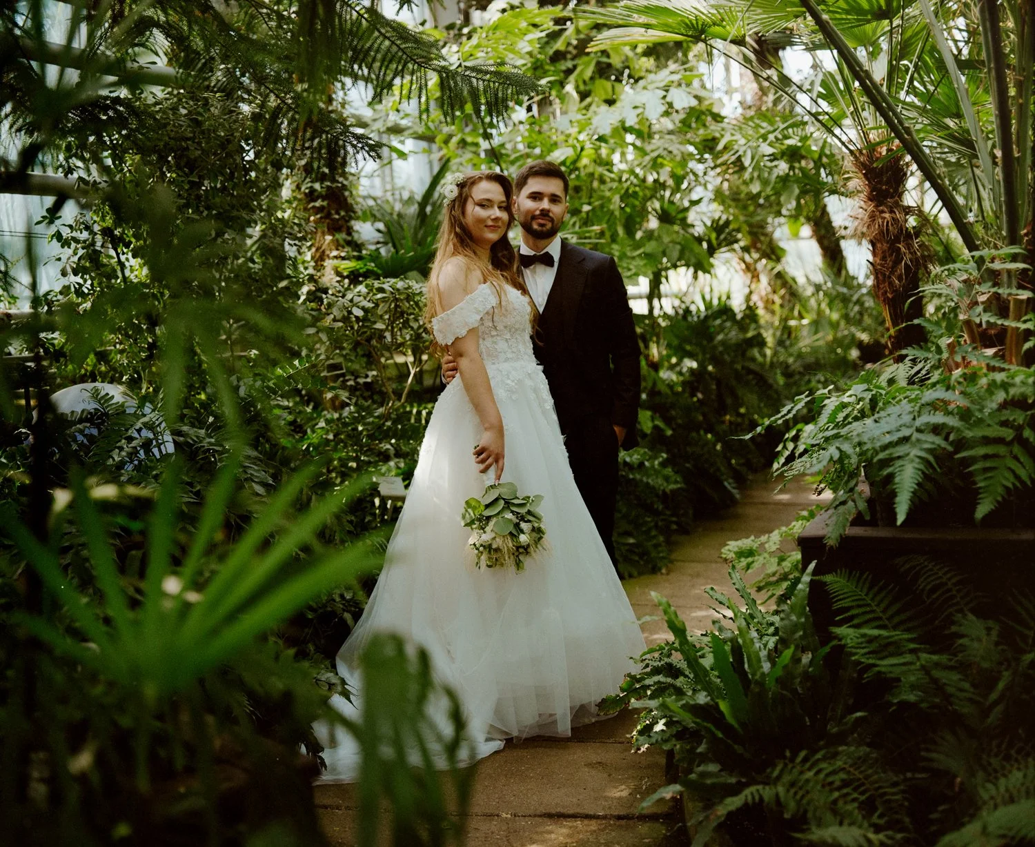 A bride and groom standing together inside a lush greenhouse. The bride wears a white off-shoulder wedding gown, holding a bouquet of greenery and white flowers. The groom wears a black tuxedo with a bow tie. They stand close, amidst abundant green plants and ferns.