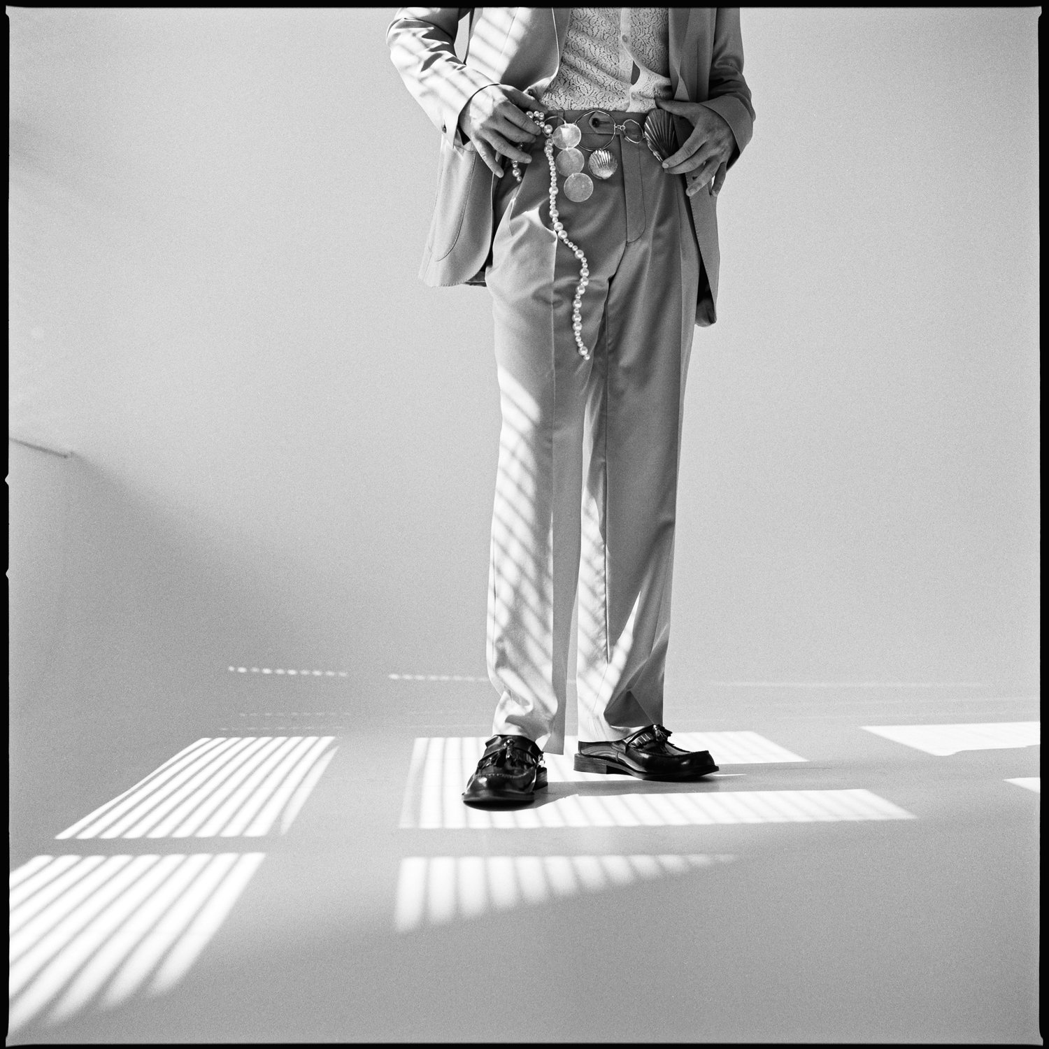 A person dressed in a suit standing on a white floor with shadows of window blinds cast across. The person's face is not visible, and they are holding sunglasses, a string of beads, and a shell in their hands.