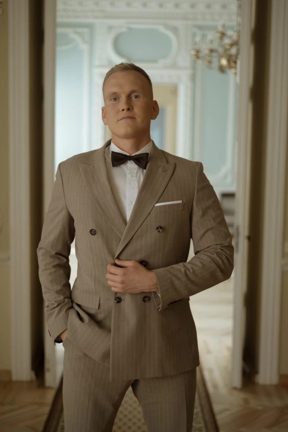 A man in a beige pinstripe suit, white shirt, and black bow tie stands in front of a mirror in an elegant room with decorative molding and a chandelier.
