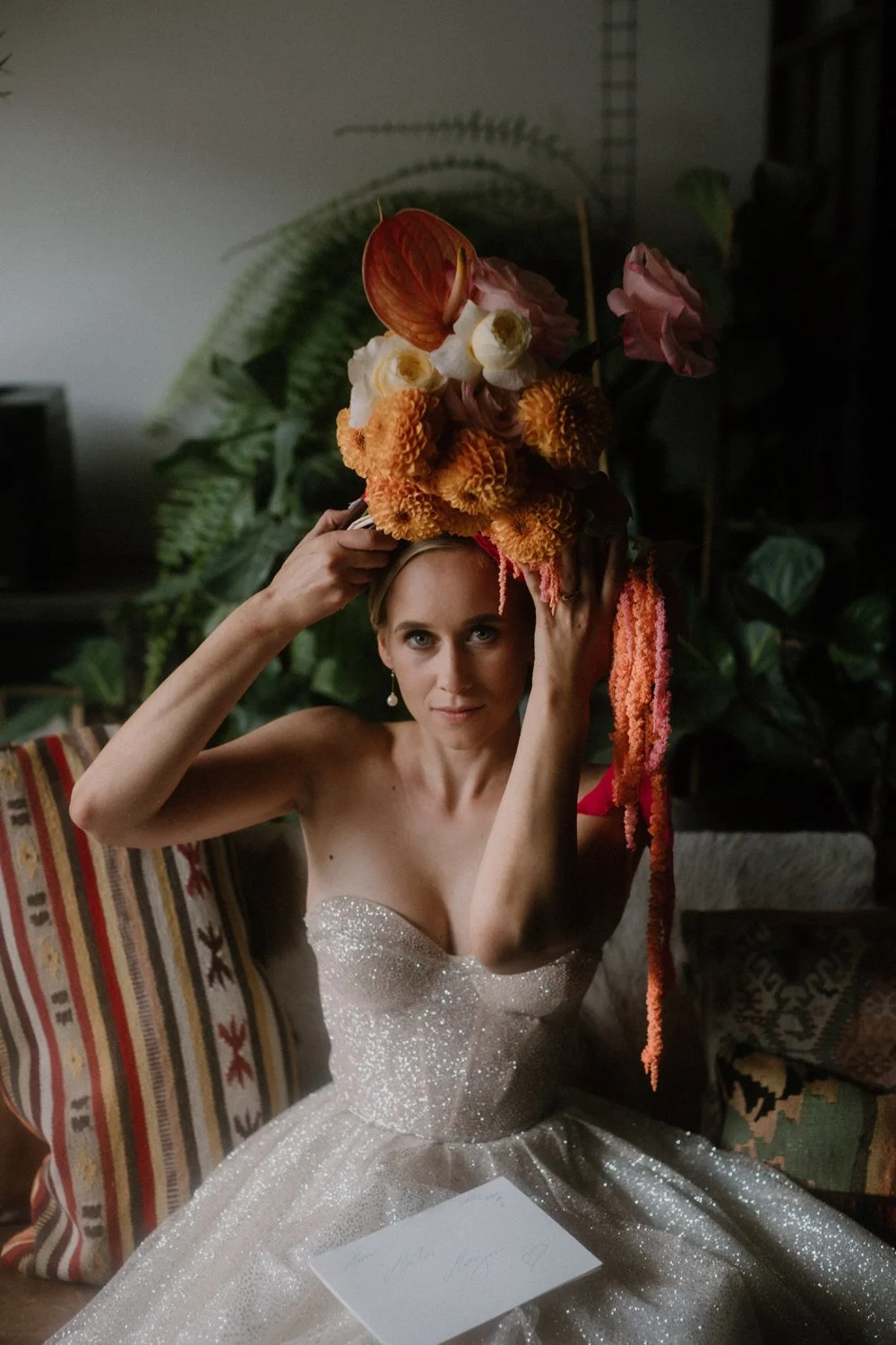 Bride in a sparkly strapless dress sitting on a couch, holding a large floral headpiece made of orange, pink, and white flowers and foliage overhead.
