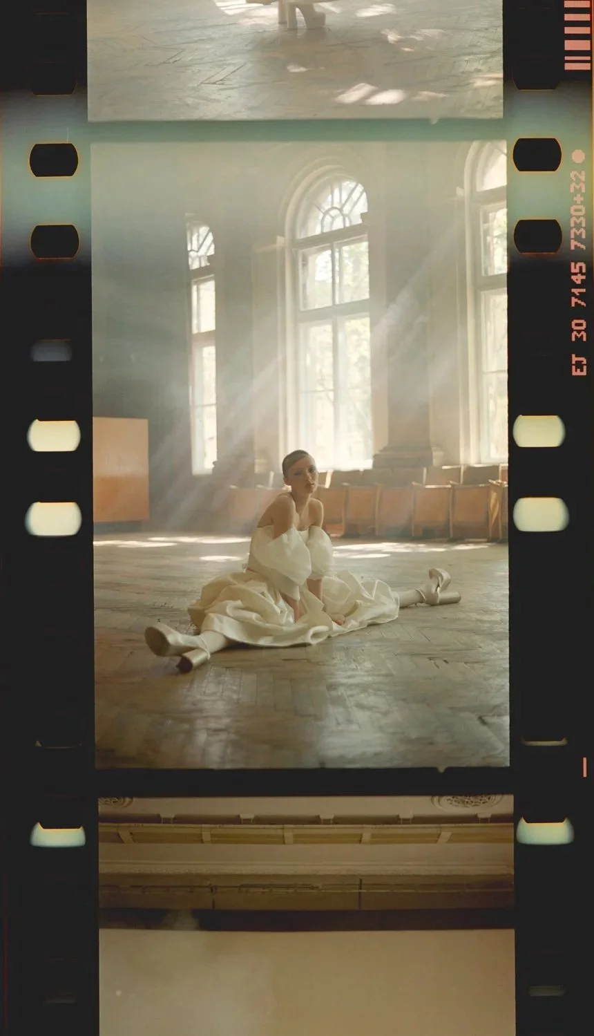 A woman in a white dress sitting on a wooden floor in a sunlit room with large arched windows.