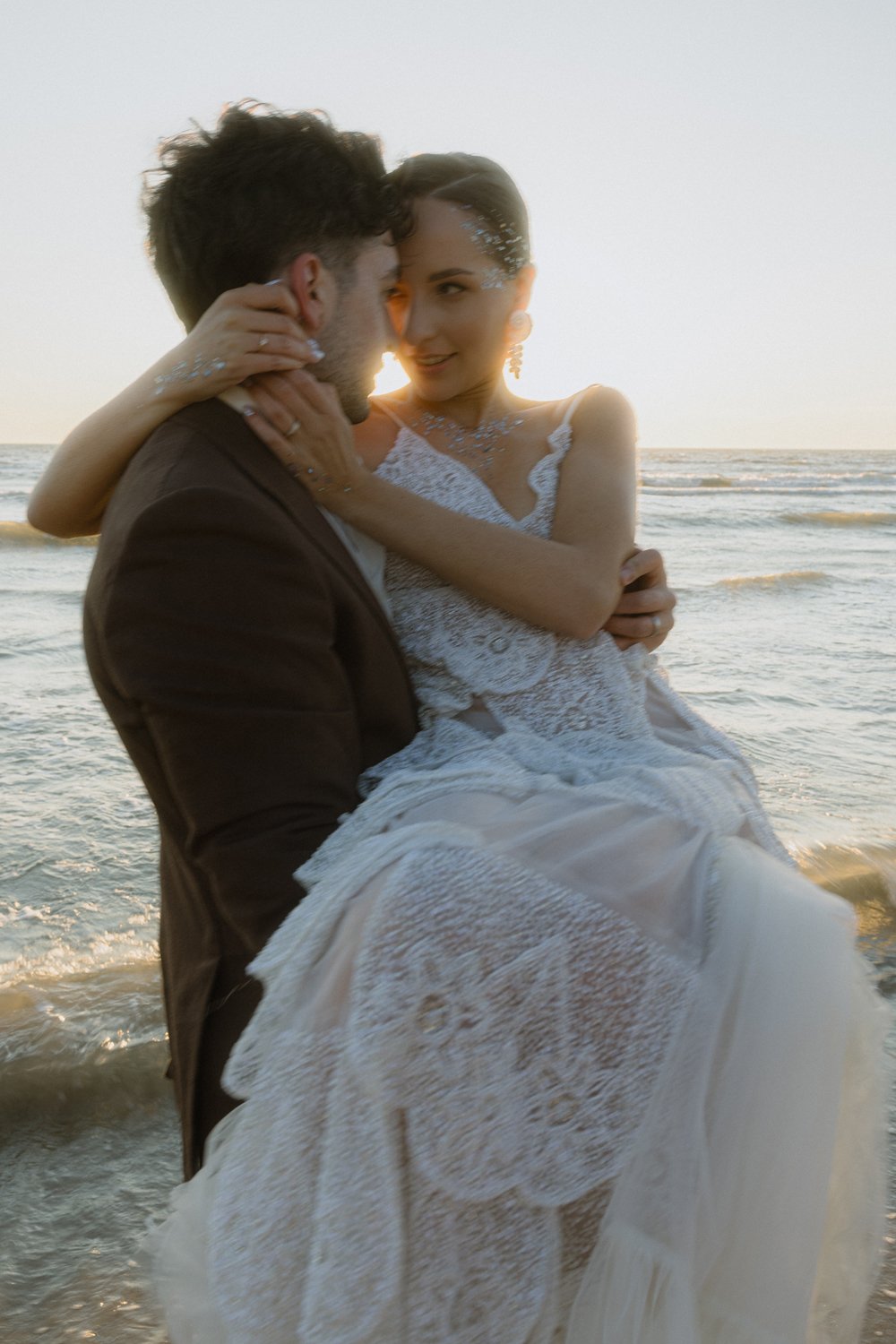 A couple in wedding attire embraces on the beach at sunset, with the ocean waves in the background.