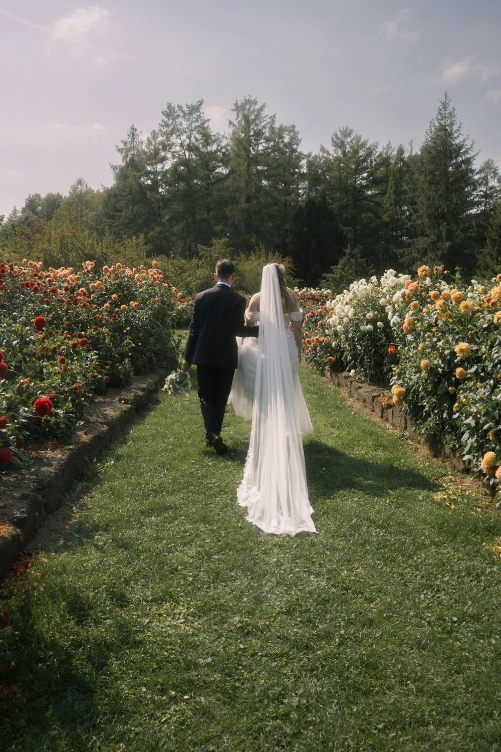 A bride and groom walking together on a grassy path through a garden of blooming flowers, with trees in the background and the bride wearing a long white dress with a veil.