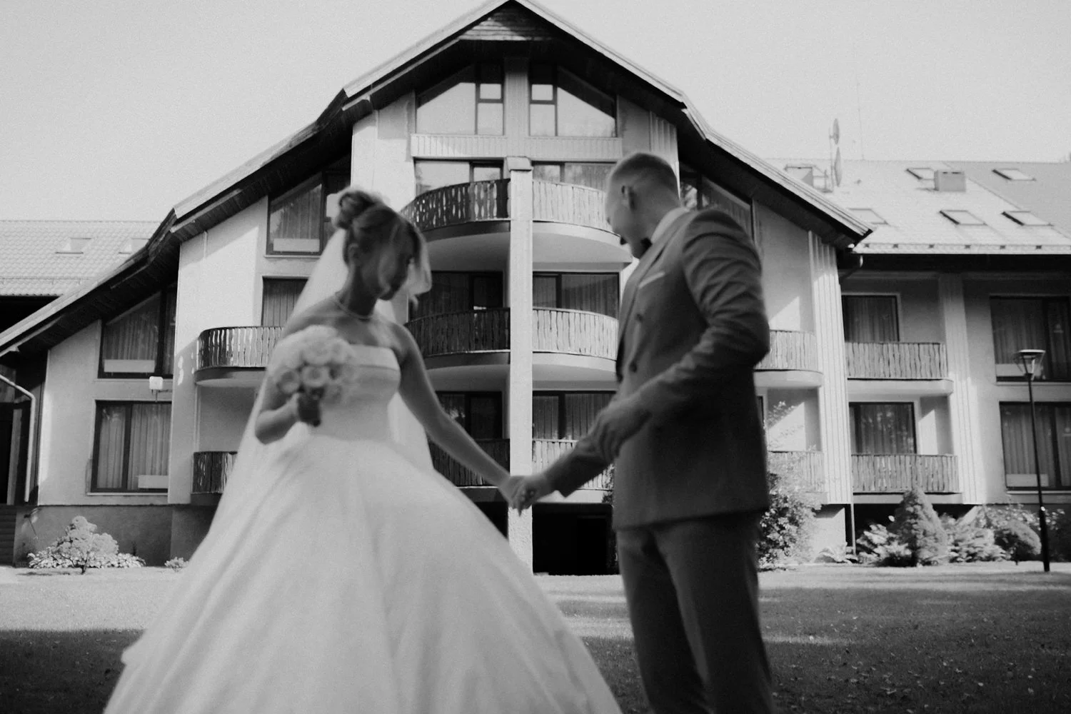 A bride in a wedding dress holding a bouquet and a groom in a suit holding hands outside in front of a modern multi-story building.
