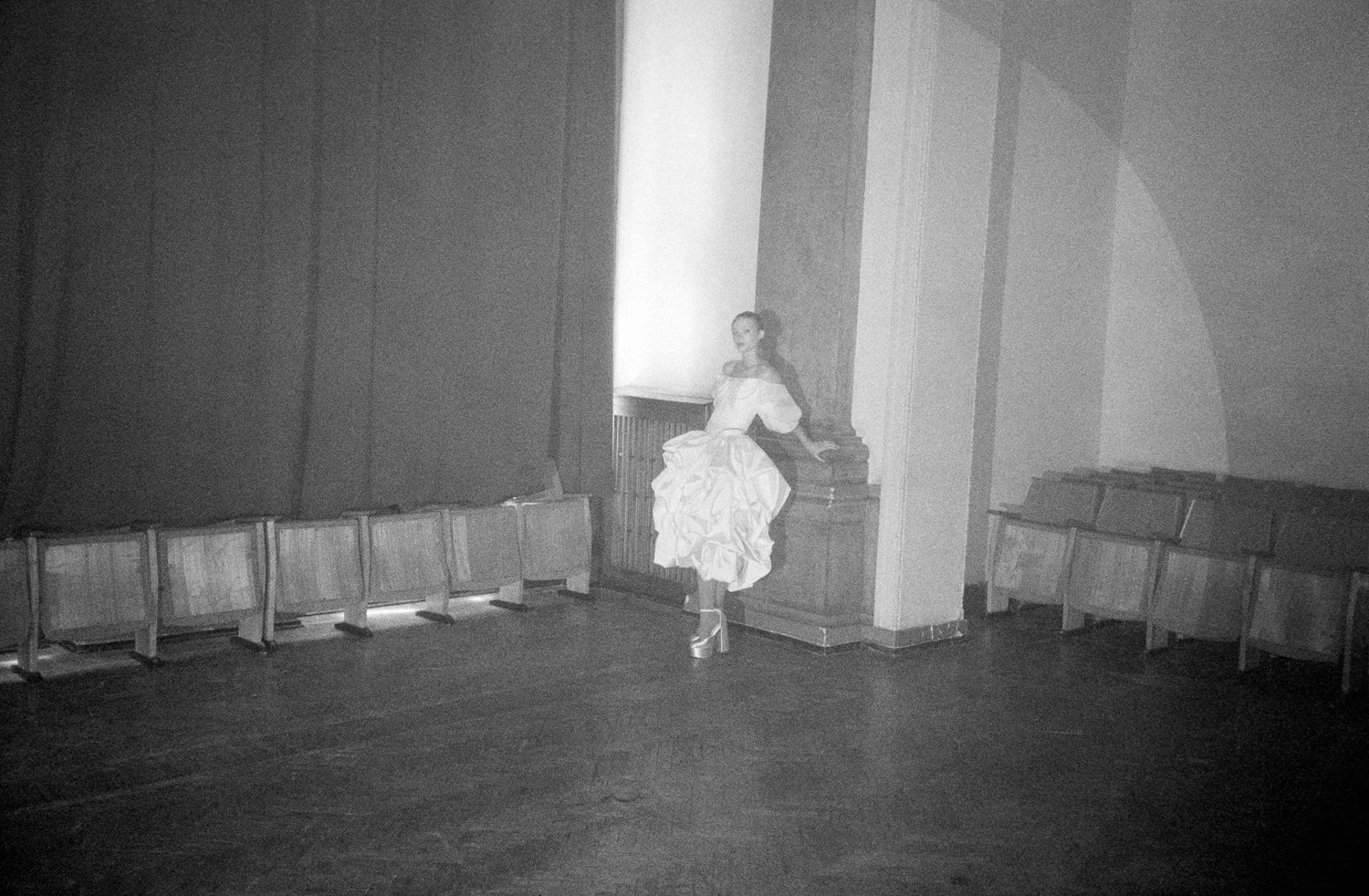 A historical black and white photograph of a woman in a long, elegant dress with a full skirt, standing on stilts in an empty room with wooden benches lining the walls, illuminated by natural light from a large window.