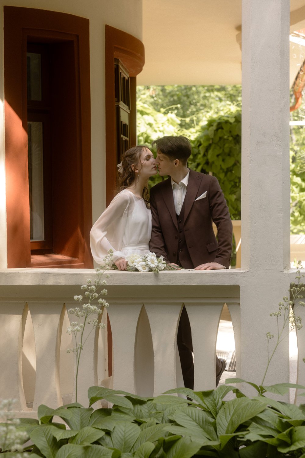 A bride and groom share a tender kiss on a porch, surrounded by greenery, during their wedding celebration.