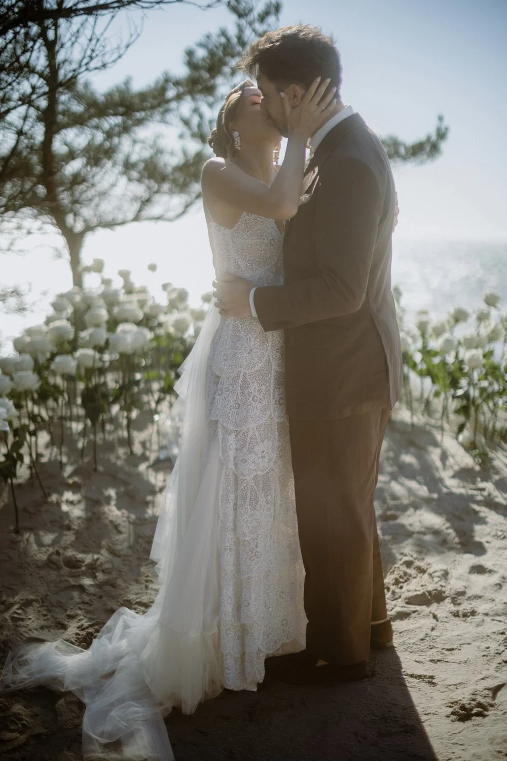A bride and groom sharing a kiss outdoors on a sunny day, with white flowers and greenery in the background.