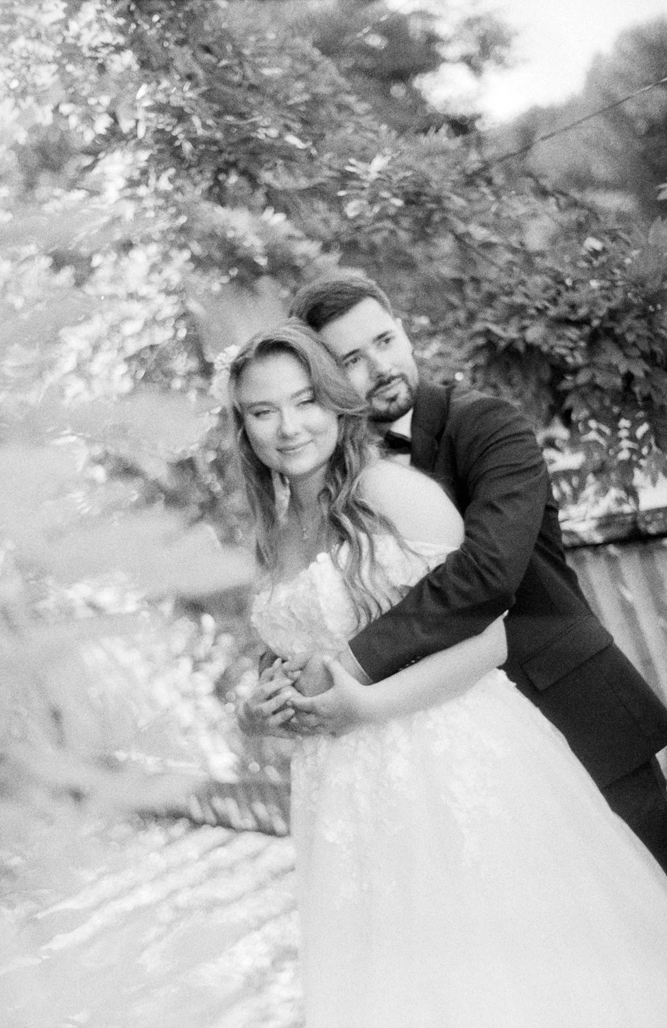A black and white photo of a bride and groom embracing outdoors with trees and a wooden fence in the background.
