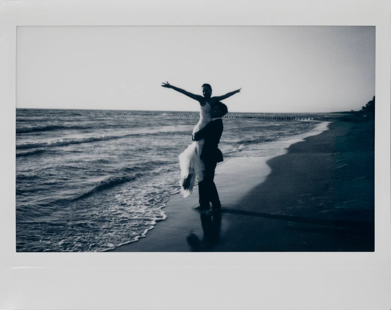 A couple on the beach, with the man holding the woman as she stretches her arms out, near the shoreline with a pier in the distance, in black and white.