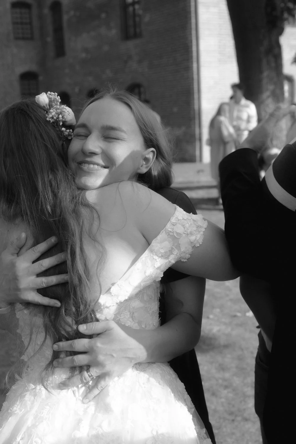 Two women hugging at a wedding, one in a floral wedding dress smiling as she hugs another person, in outdoor setting with a brick building and trees in the background.