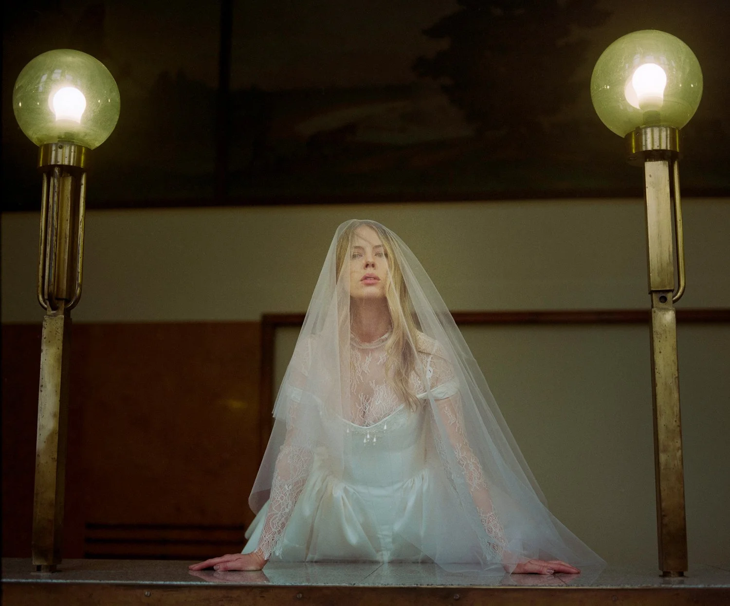 A woman dressed as a bride in a white wedding gown and veil, kneeling on a table with her hands on the surface, between two vintage-style lamps with greenish glass globes.