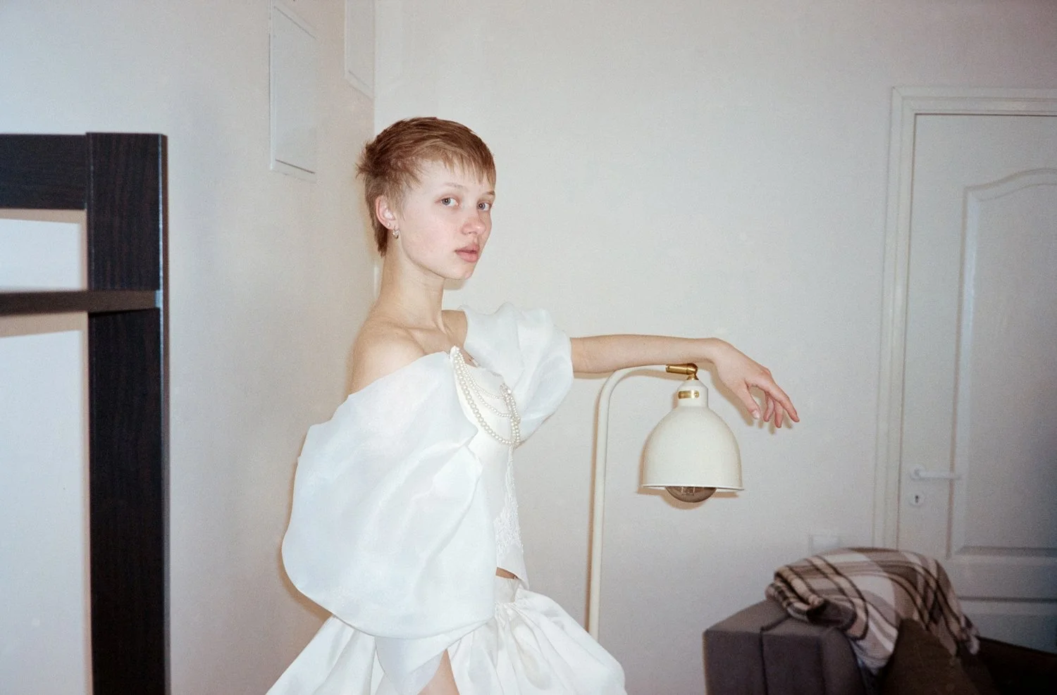 A young woman with short hair, wearing a white dress and pearl necklace, poses indoors next to a wall lamp, with minimal furniture and a plaid cloth covering on a piece of furniture.