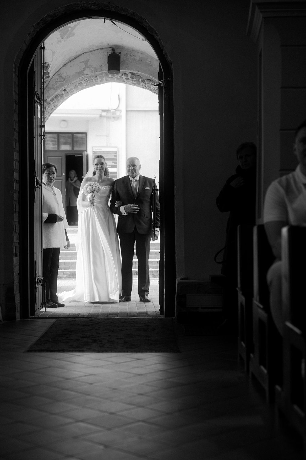 Black and white photo of a bride in a wedding gown walking down the aisle with a man in a suit, accompanied by two women, possibly her family members, at the entrance of a building.