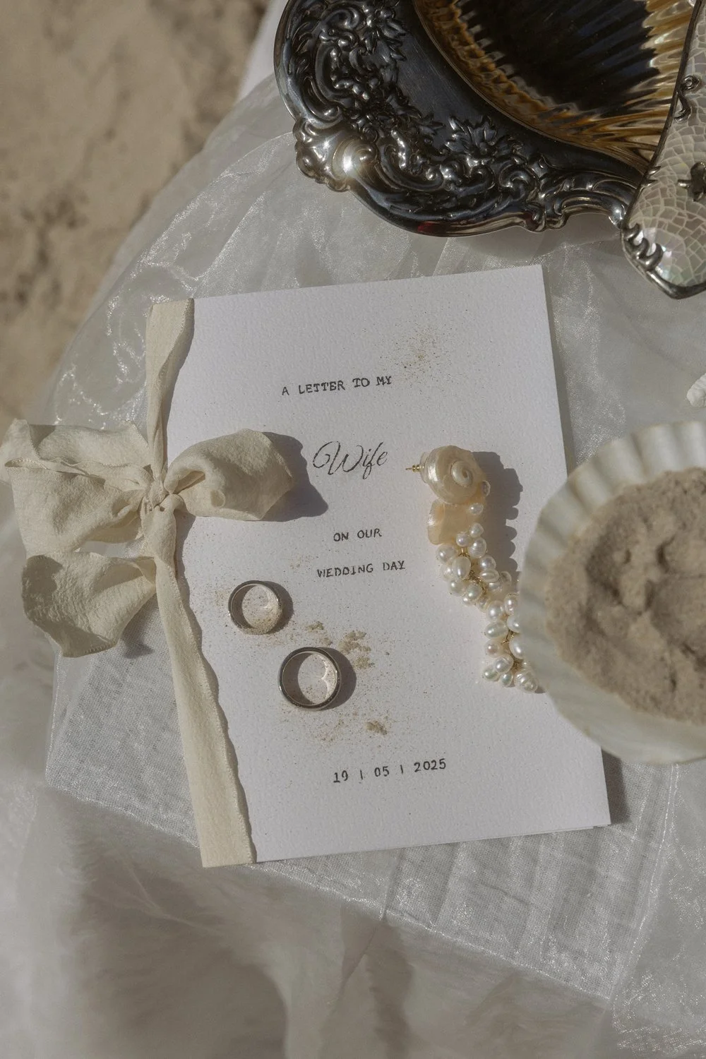 A wedding invitation card with the text 'A letter to my Wife on our wedding day 19 05 2025,' accompanied by two wedding rings, a pearl ring with a large pearl and smaller pearls, a cream-colored fabric bow, and decorative silver and glass items on a 