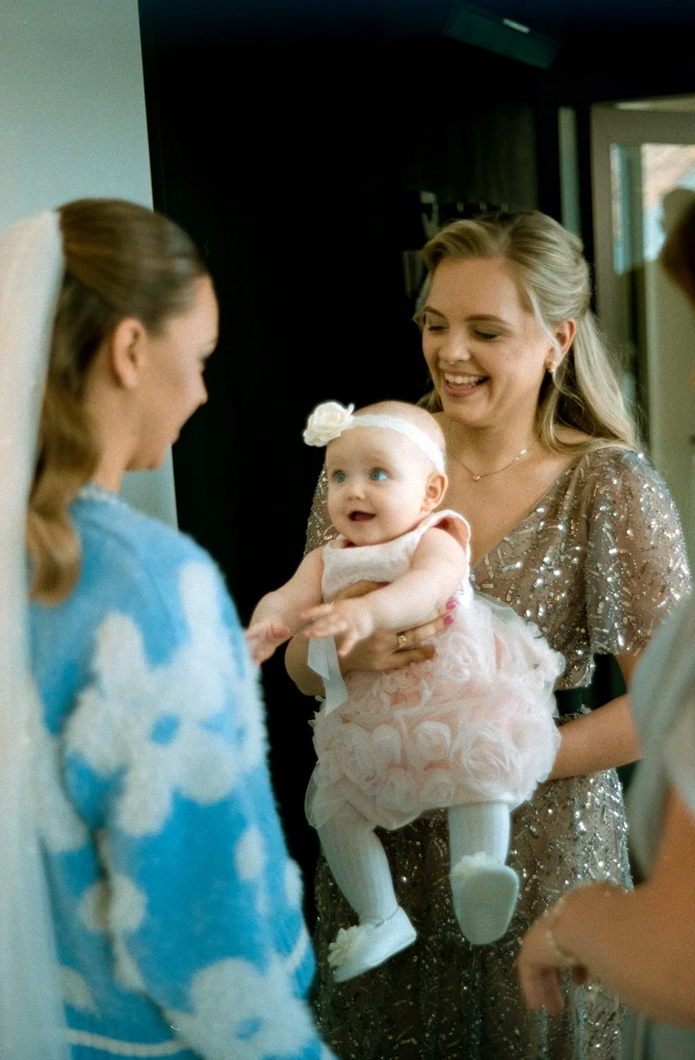 A woman holding a smiling baby girl in a pink dress and headband, surrounded by friends or family in a brightly lit indoor space.