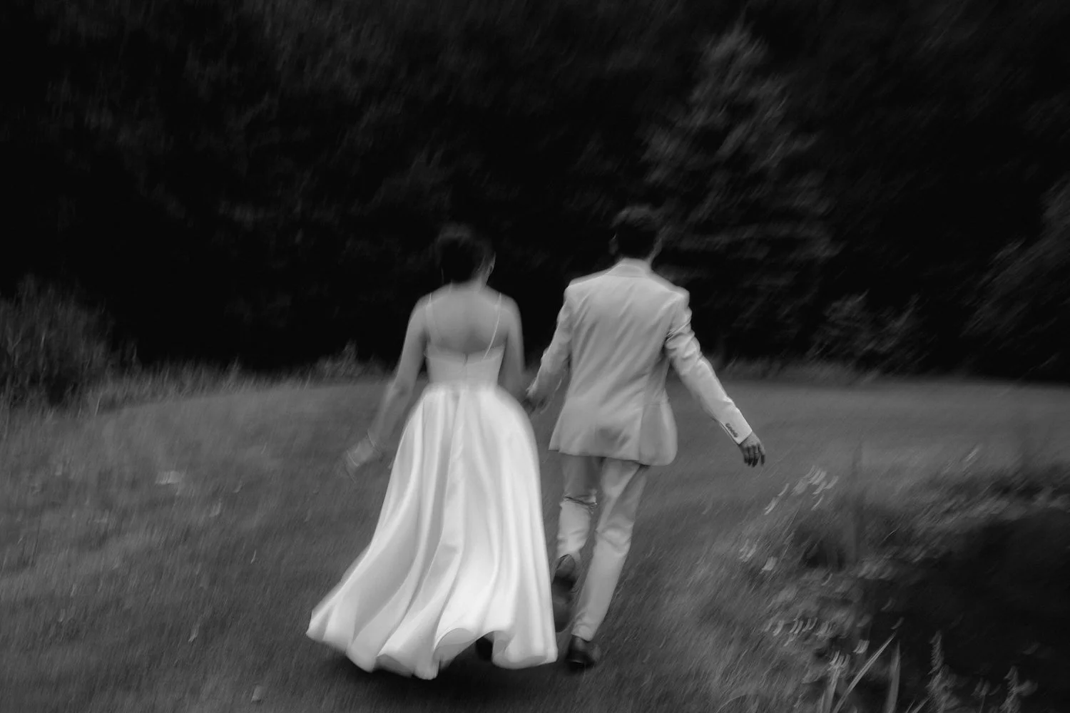 A bride and groom walking hand in hand on a grassy field, dressed in wedding attire, with a blurry dark sky or wooded area in the background, captured in black and white.