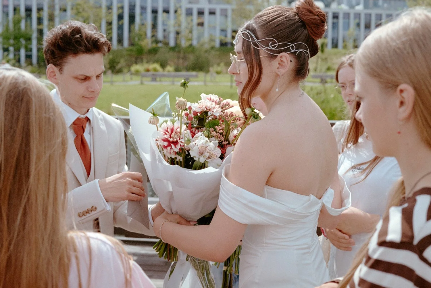 A bride holding a bouquet of pink and white flowers during a wedding ceremony, surrounded by bridesmaids and groomsmen outside.
