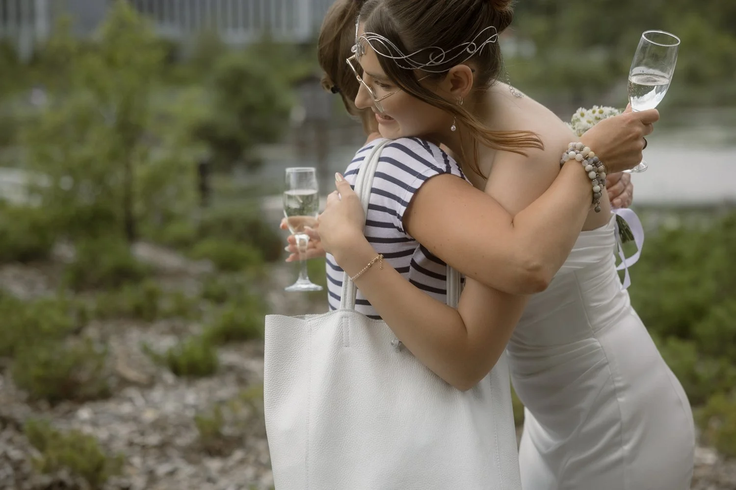 Two women hugging outdoors, each holding a champagne flute, with a blurred natural background.