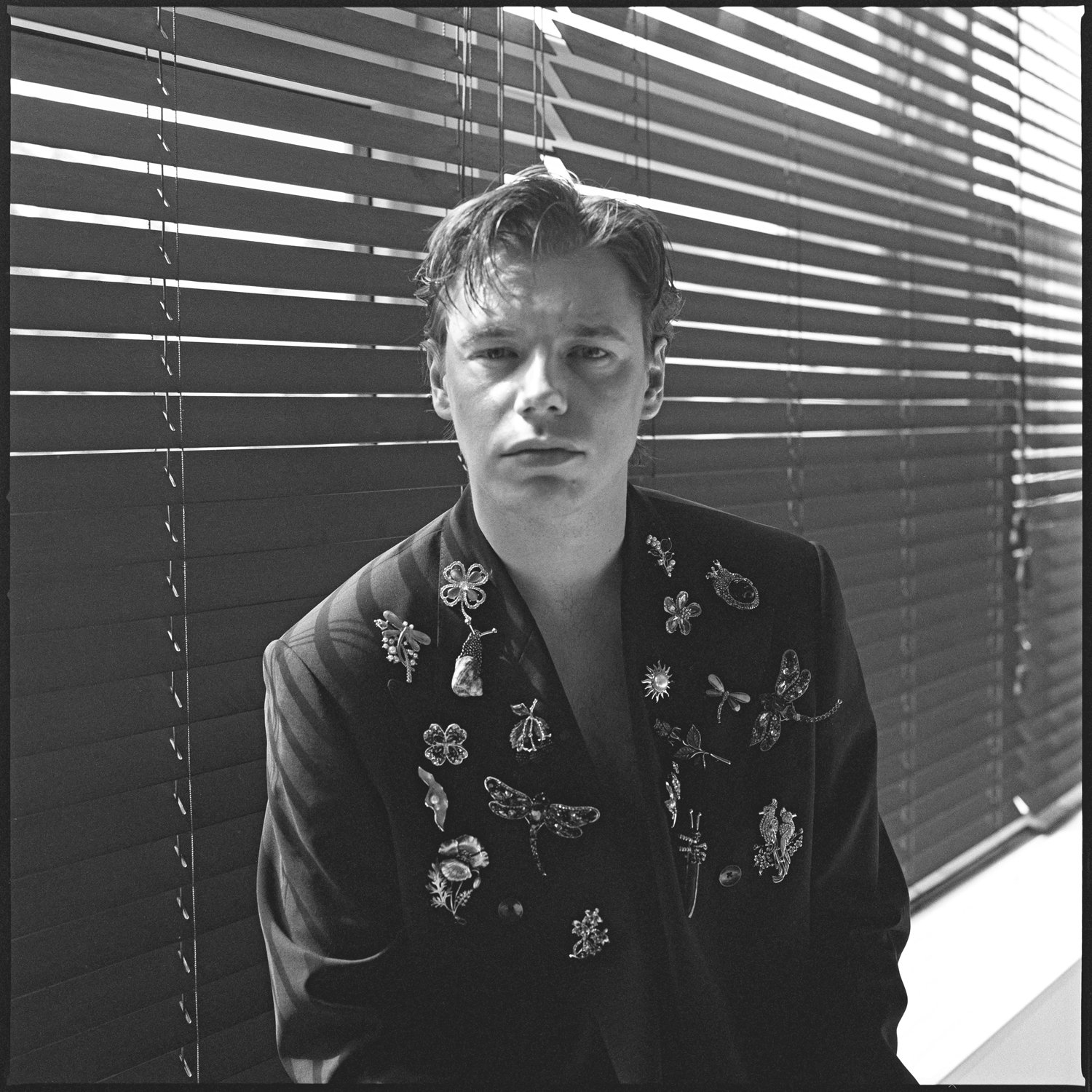 Black and white photo of a young man with styled hair, wearing a dark blazer adorned with various pins and decorative pieces, standing in front of closed window blinds.