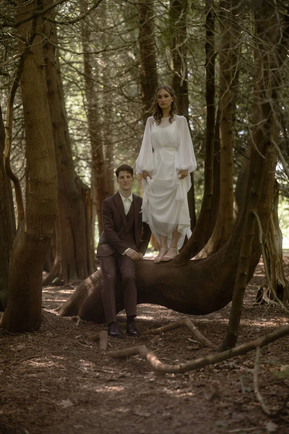 A woman in a white dress walking on a tree branch in a forest with a man in a dark suit sitting on the branch below her.