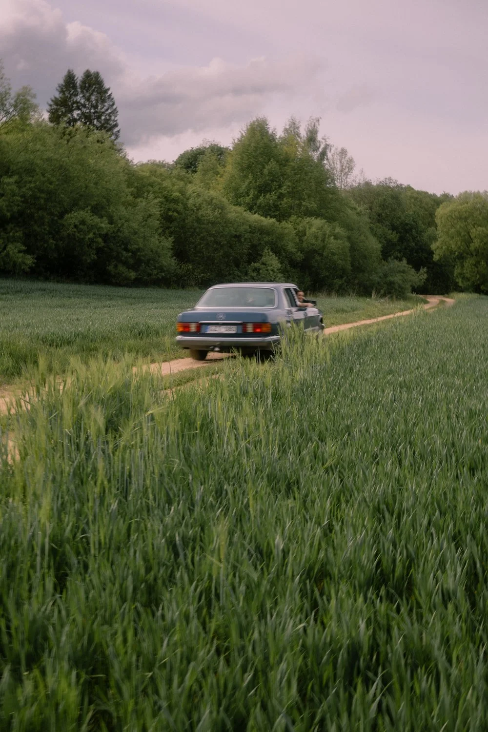 A vintage blue car driving on a dirt road through green fields with trees in the background under a cloudy sky.