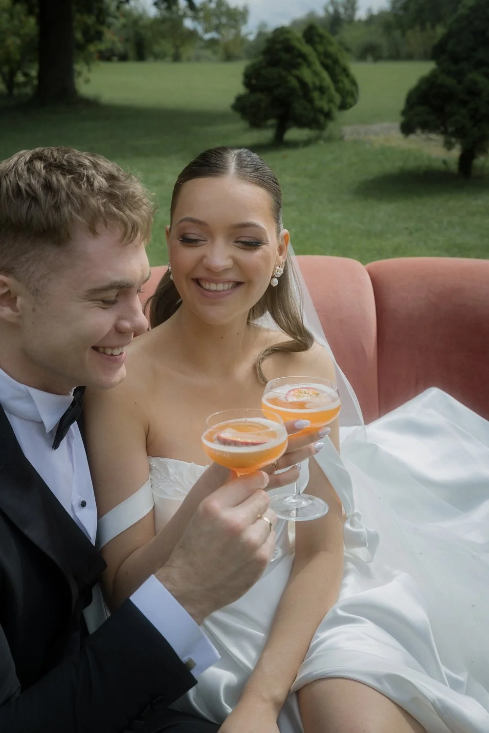 A happy bride and groom in wedding attire sitting on a pink couch outdoors, holding glasses with pink cocktails garnished with citrus slices, smiling at each other.