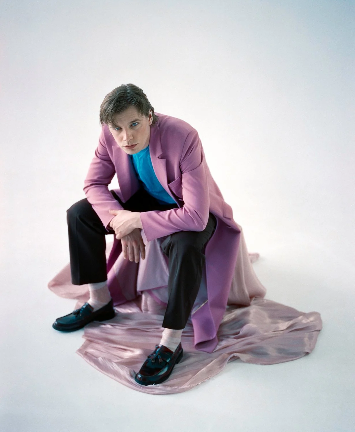 A man wearing a pink coat, black pants, and black shoes, sitting on a pink fabric-covered platform against a white background, with a serious expression and looking at the camera.