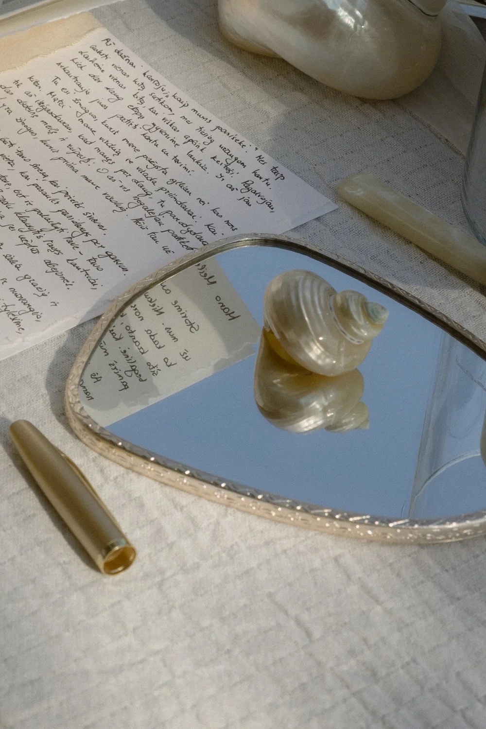 A decorative table setup featuring a reflective silver heart-shaped mirror with a seashell ornament on top. There is handwritten paper with cursive writing, a gold-colored pen, and a beige cloth backdrop, creating an elegant, beach-inspired aesthetic