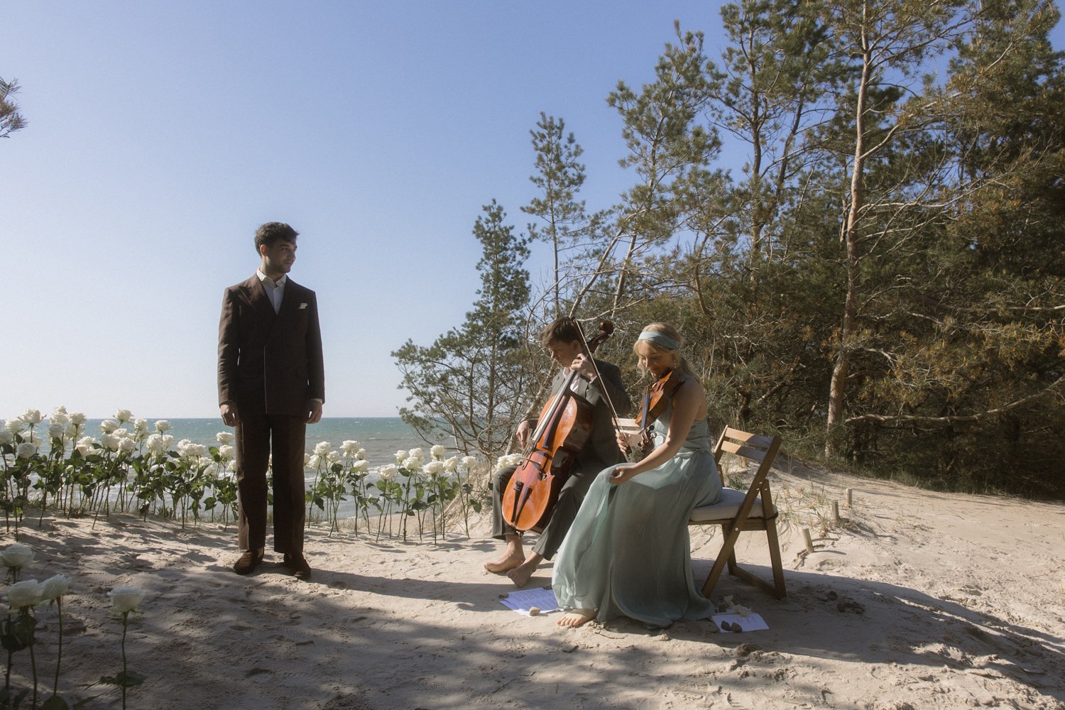 Two musicians playing cello and violin while a man in a suit stands nearby on a sandy beach with flowers and trees, against a backdrop of ocean and blue sky.