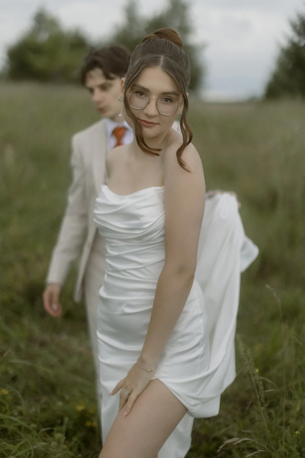 A young woman in a white dress and glasses poses outdoors in a grassy field, with a young man in a suit blurred in the background.