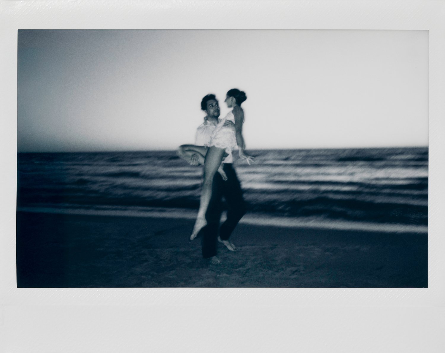 A black-and-white photo of a man holding a woman in his arms on a beach with ocean waves in the background.