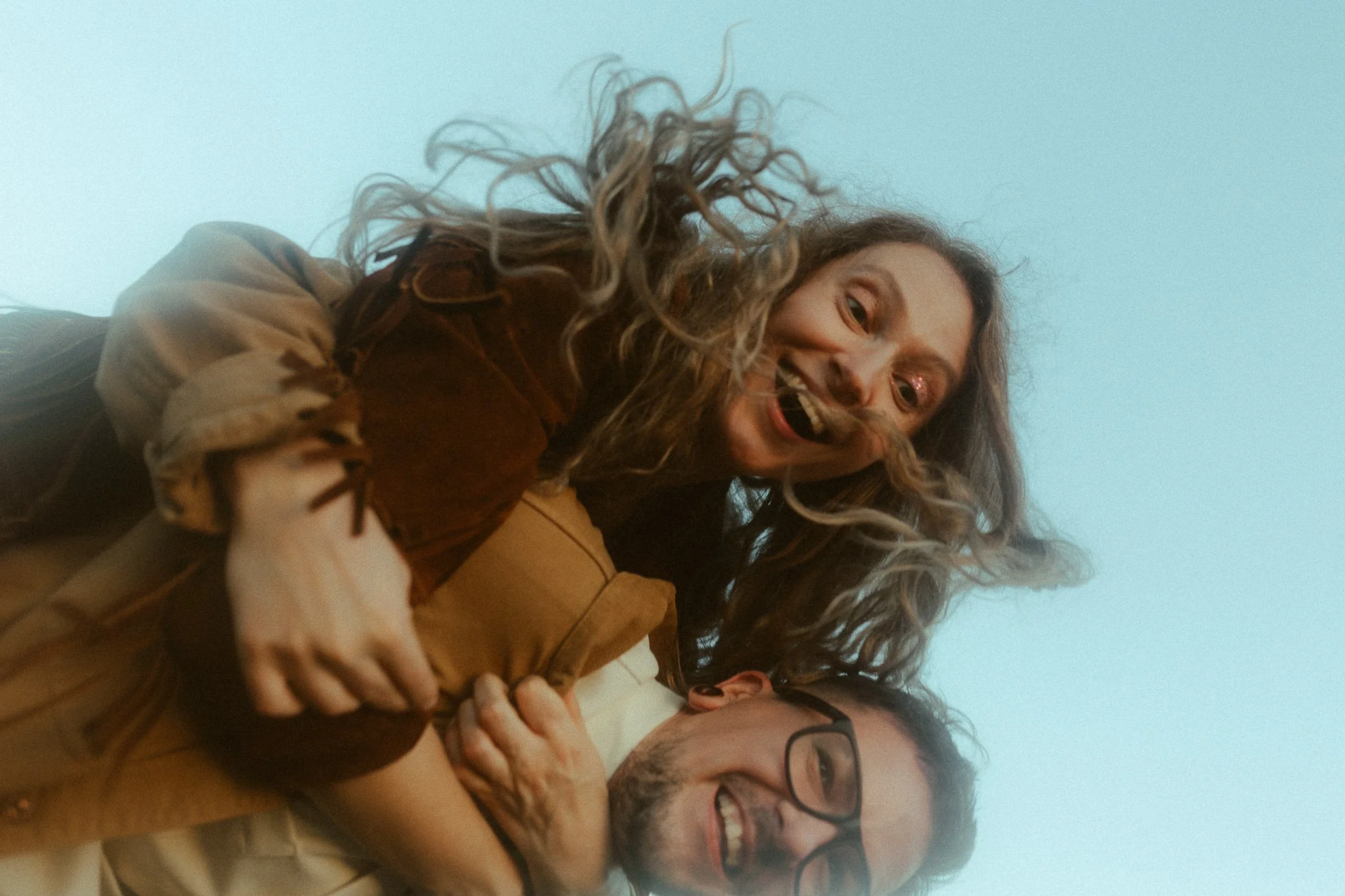 A happy couple, a woman with curly hair and a man with glasses, smiling and embracing each other against a clear blue sky.
